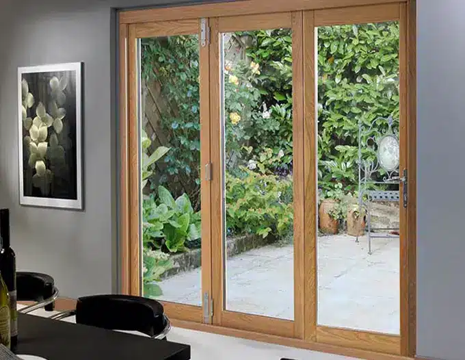 A set of three-panel bi-fold patio doors with warm oak wood frames opened to reveal a lush green backyard garden with outdoor seating on a stone patio. The doors feature full-length glass panels that flood the interior room with natural light, with a framed black-and-white floral print visible on the adjacent gray wall.
