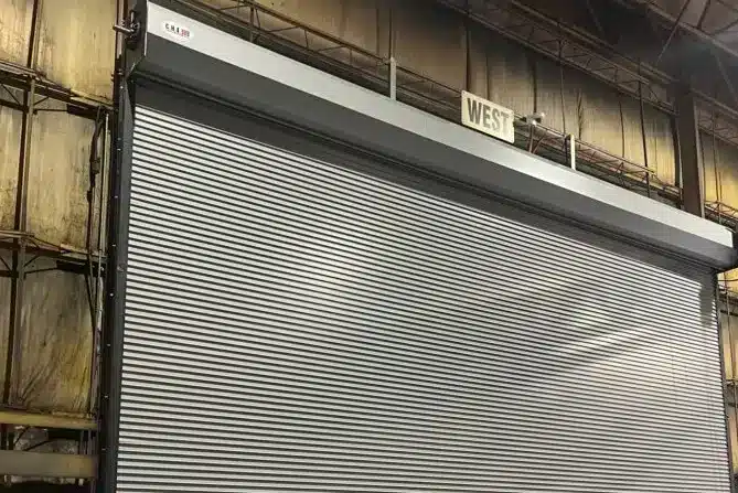 A large commercial rolling steel door fully closed inside an industrial warehouse bay, showing horizontal ribbed metal slats with a slight reflective sheen under overhead lighting. The door spans the full width of the loading area, with a metal housing unit mounted above the opening and weathered concrete and steel framing visible on either side, typical of multi-bay warehouse installations in commercial settings.