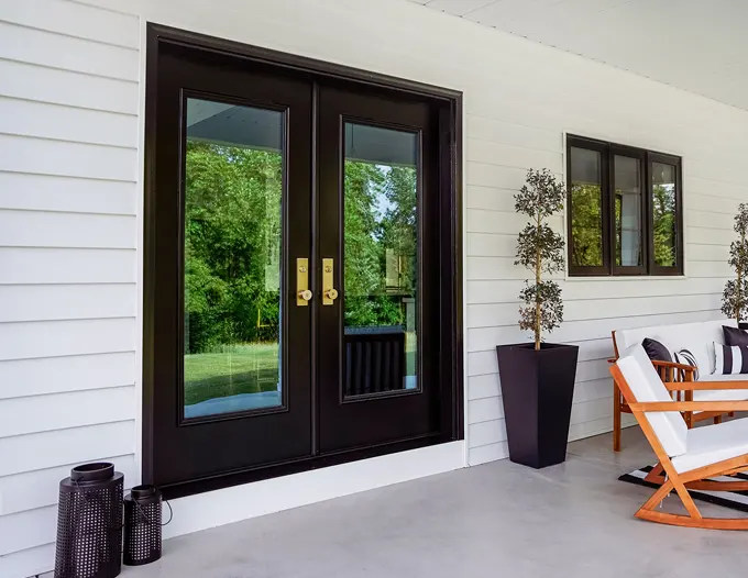 A modern covered porch featuring elegant black French double doors with large glass panels and brass handles, flanked by a dark planter with a topiary tree and decorative black vases, with outdoor wooden furniture visible to the right. The doors open to a lush green yard visible through the glass, set against a clean white horizontal lap siding exterior. This style of front entry door installation reflects the premium residential door options available for Las Vegas homes.