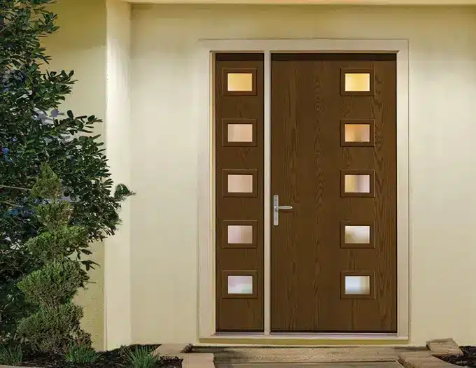A modern front entry door with a rich brown wood-grain finish featuring two vertical columns of square frosted glass inserts, flanked by a narrow sidelight panel with matching glass cutouts. The door is set within a clean cream-colored stucco exterior typical of Las Vegas desert homes, with a silver lever handle and a small landscaped area with green shrubs to the left. The contemporary design balances privacy and natural light, showcasing the type of custom front door installation suited for desert residential properties.