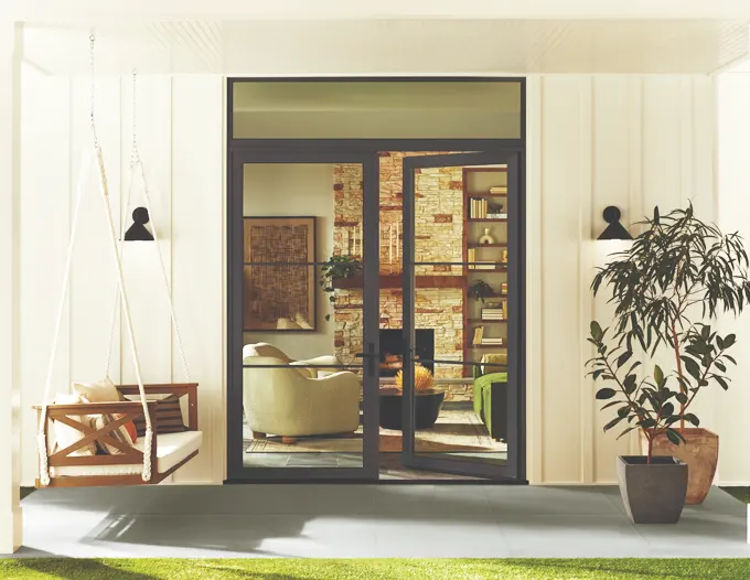 A modern home exterior featuring large black-framed sliding glass patio doors open to reveal a warmly lit living room with bookshelves and comfortable seating inside. The entry is flanked by white vertical siding, a wooden bench swing on the left, a tall potted plant on the right, and two black wall-mounted sconce lights, all set on a sunlit stone patio with green groundcover.