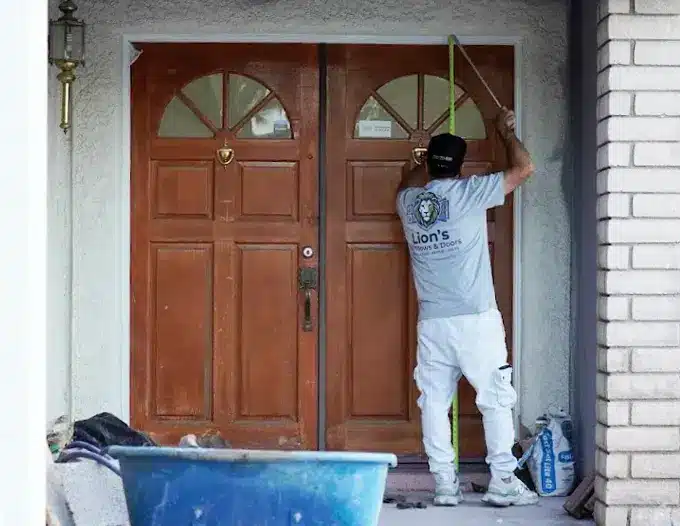 A technician from Lion's crew wearing a gray branded shirt, white pants, and a dark cap carefully works on the upper trim of a set of large double wooden front entry doors with arched transom windows, preparing the entryway for finishing or installation. Painting supplies including a blue bin and bottles sit nearby on the porch of a white brick residential home, reflecting the hands-on, same-crew approach used for every front door installation project.