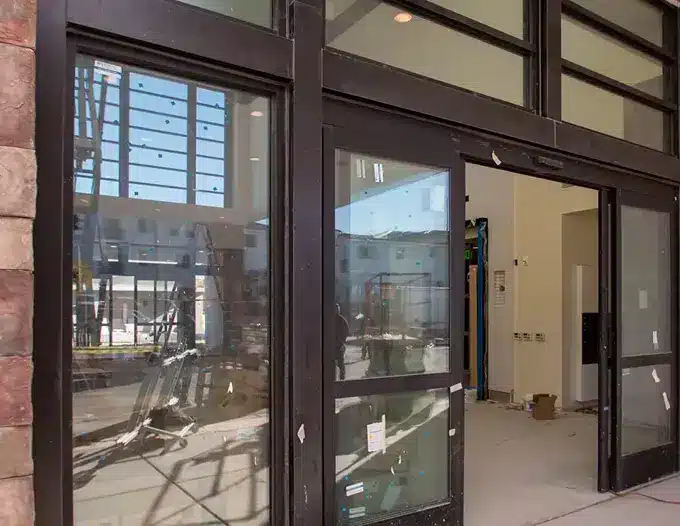A dark bronze aluminum storefront door and window system during commercial installation in Las Vegas, showing large glass panels set in metal frames with construction materials visible on the ground and building interior partially visible through the glass. The entry system features a single swing door flanked by fixed sidelight panels and transom windows above, typical of a retail or commercial storefront setup awaiting final completion.
