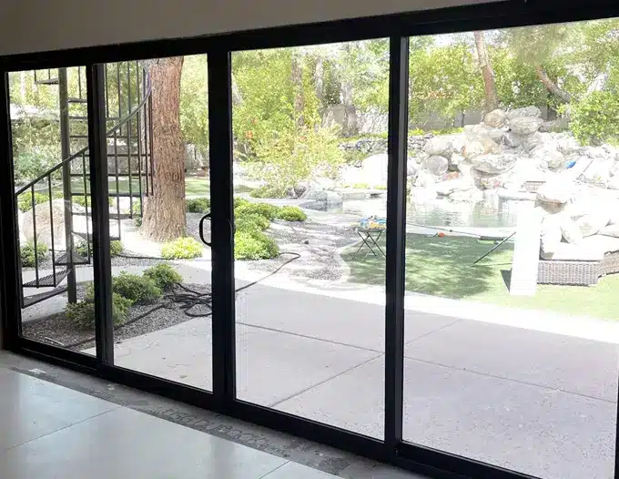Large black-framed multi-panel sliding glass patio door system viewed from inside a home, showing a landscaped backyard with mature trees, decorative boulders, a manicured lawn, and a concrete patio area. The floor-to-ceiling glass panels feature slim aluminum frames in a modern profile, allowing abundant natural light while providing a wide view of the outdoor living space. The clean, contemporary design and desert-compatible landscaping reflect a Las Vegas residential installation suited to indoor-outdoor desert living.