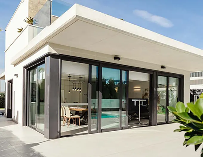 A modern residential building featuring fully open large-format sliding glass doors with slim dark aluminum frames, revealing an indoor living and dining area that flows seamlessly into the outdoor patio space. The flat-roofed contemporary architecture showcases floor-to-ceiling glazing panels folded open in an accordion configuration, demonstrating the type of premium sliding door installation suited for desert climates. Green landscaping accents the foreground while the bright Las Vegas sky fills the background.